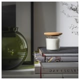 Small round glass jar with white candle and cork lid sits atop books next to green vase.