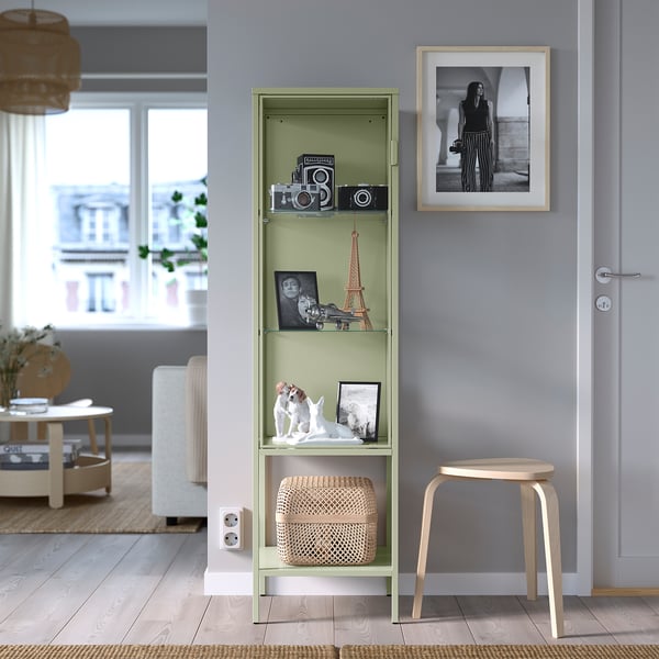 Green glass-doored cabinet with metal shelves displays items, stands near door between stool and couch, adjustable feet.