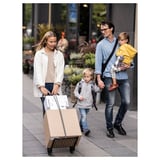 A family walks on a city street. The woman pulls a beige RULLEBÖR cart with purchases. The man holds a child and a teddy bear.