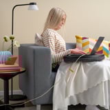 Woman in striped shirt works on laptop at table with lamp and flowers.