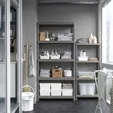 Gray storage shelf with multiple levels holding clothes and supplies against wall, next to mop and bucket.