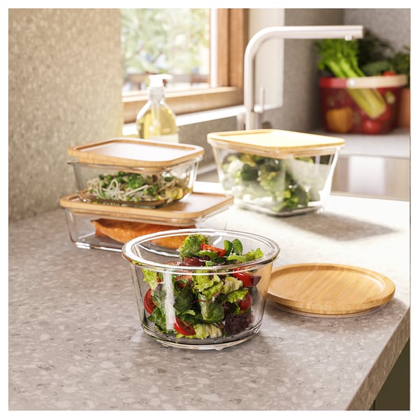 Three glass containers with wooden lids hold fresh salads on a beige countertop, with one in the foreground.