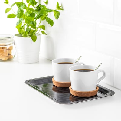 Two white coffee cups with saucers on a metallic tray. Green spoons rest in cups. Tray sits on a white countertop near a plant and cookies.