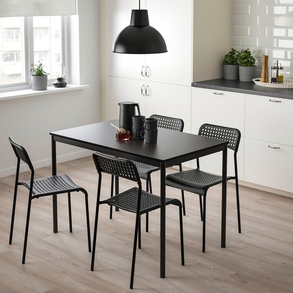 Black sandberg dining table with 4 stackable chairs, kettle, mugs, near kitchen cabinet, under pendant light, on light wood floor.