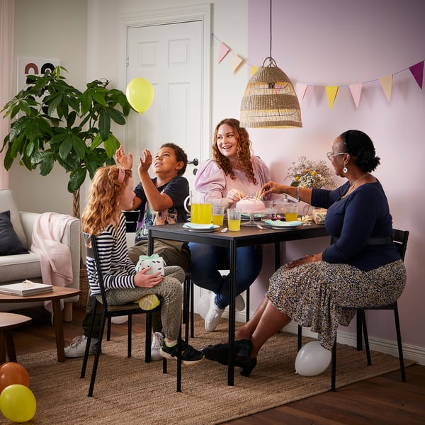 Family celebrates at black sansberg table with yellow drinks; adult holds cake, child plays with balloon.