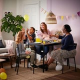 Family celebrates at black sansberg table with yellow drinks; adult holds cake, child plays with balloon.