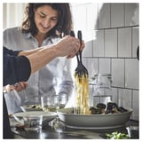 Woman serving noodles from skillet to bowl with SANDSKÄDDA gray tableware and glasses.