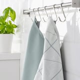 Two light blue towels hang on silver hooks under a white shelf. A green plant sits on the left.