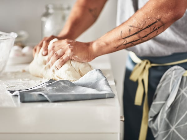A person kneads dough on a floured counter, wearing a gray apron.