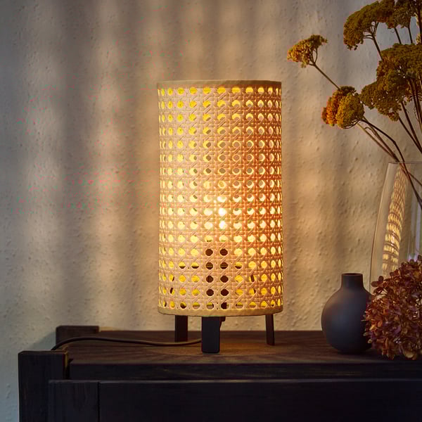 A SAXHYTTAN lamp with a perforated paper shade, casting warm light. It sits on a wooden surface with dried flowers in a vase beside it.