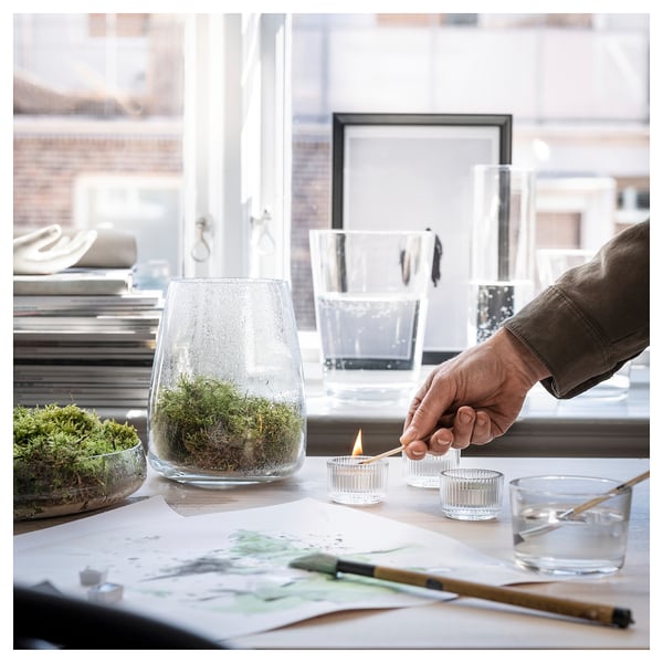 Hand lights white candle near clear glass vase with greenery on wooden table by window.