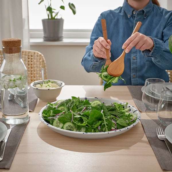 Person tossing salad into bowl with tongs. Table set with glassware, cutlery, and sauce.