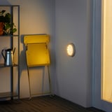 Motion-activated SJÖNÖT light on wall, illuminating corner with chair and plant shelf.