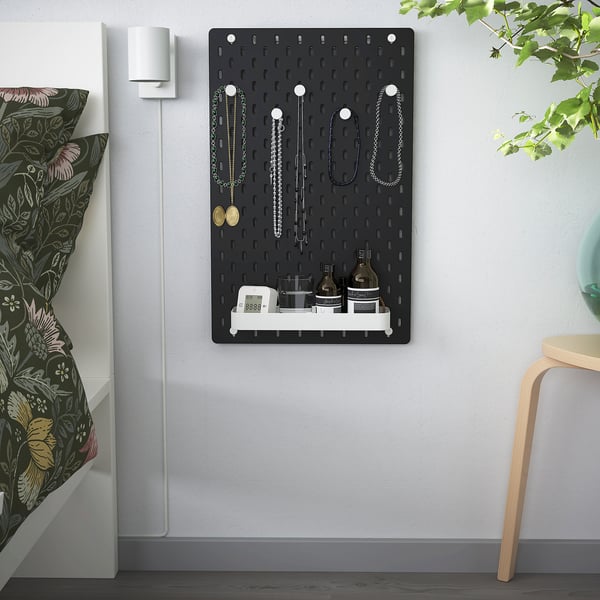Black SKÅDIS pegboard with accessories, beside bed with floral pillow and wooden chair.