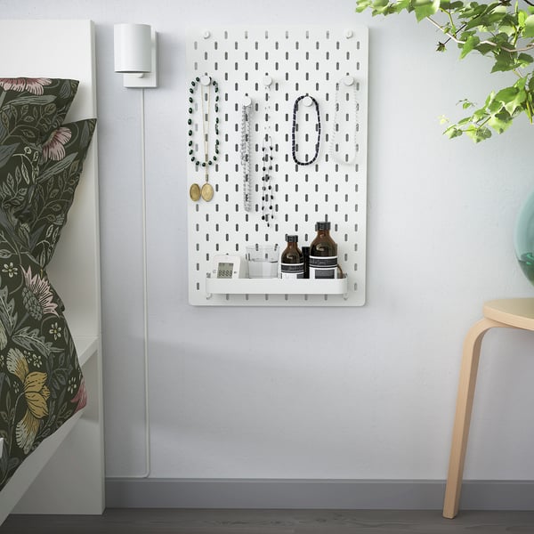 SKÅDIS pegboard with jewelry, bottles, clock. Beside bed with floral pillows and chair.
