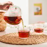 Hand pours tea from glass teapot into cup on woven mat. Teapot has built-in infuser and heat-resistant glass.