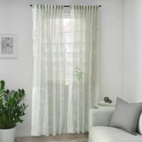 Sheer SKÄREFLY curtains in cream with floral design cover large window, flanked by houseplant and grey cushion on chair.