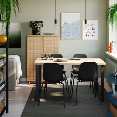 Modern dining area with SKÅLSTA table and black chairs. Light wood table, dark metal legs. Bright art, plants, wicker cabinet. Cozy, casual setting.