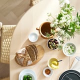 Round light wood dining table with extension, surrounded by wicker chairs, set for a meal.