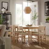 Modern dining room with light wood table and chairs, hanging lamp, plant, and bookshelf.