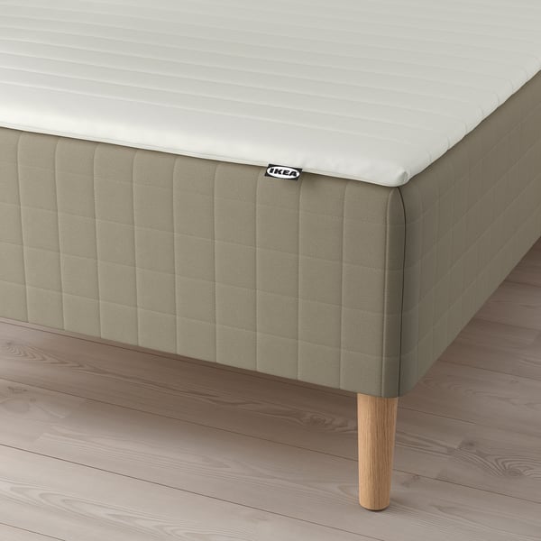 A modern bed setup with SKÅRER mattress on a wooden base, featuring a beige cover, foam topper, and wooden legs from IKEA.