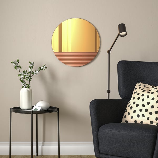 Rose-gold mirror in living room beside dark grey armchair with pillow, black side table, white vase, flowers, sphere.