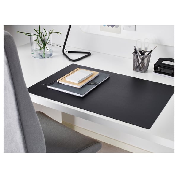 Modern workspace with black SKRUTT desk pad, notebooks, pen holder, and small plant. Pad covers desk, has smooth surface.