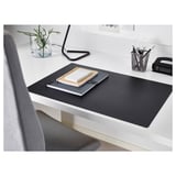 Modern workspace with black SKRUTT desk pad, notebooks, pen holder, and small plant. Pad covers desk, has smooth surface.