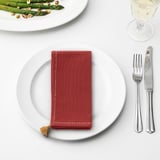 SLÄTTIKA red napkin, small with tassel, on plate, knife, fork, glass, asparagus nearby.