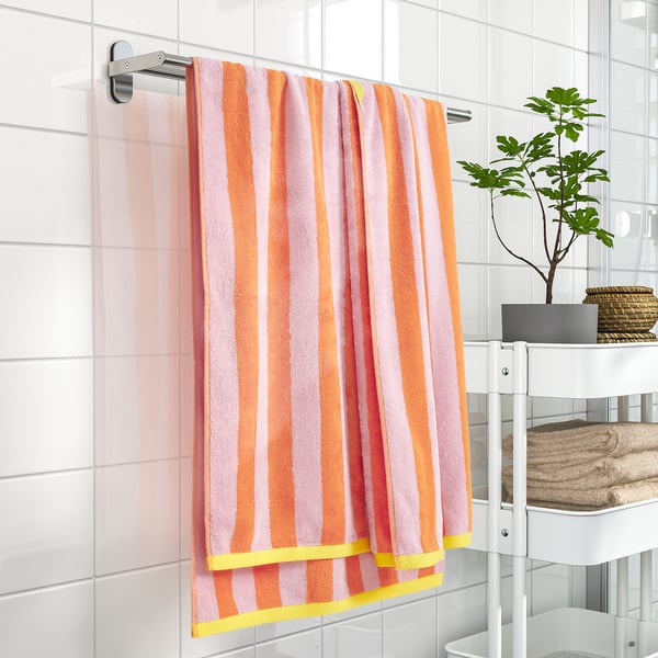Striped SLÅNHÖSTMAL towel hangs on wall rack, bright colors, 100% cotton, framed by tiles, next to small plant and folded towels on shelf.