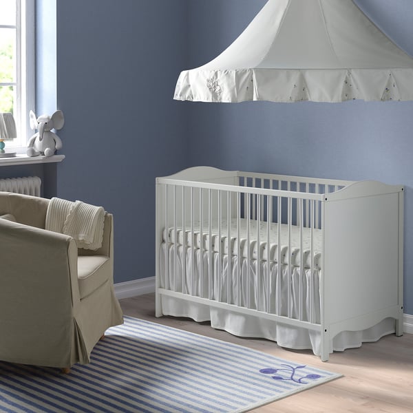 A SMÅGÖRA white cot with bevelled edges in a blue nursery, next to a grey armchair and under a white canopy, with a blue-striped rug.