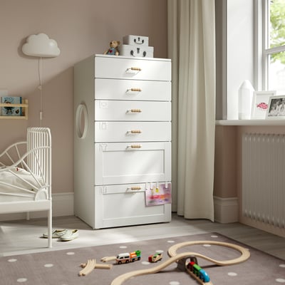 Kids room with white SMÅSTAD chest, 7 drawers, wooden handles. Near white bed, window with beige curtains, toys, and rug with polka dots.