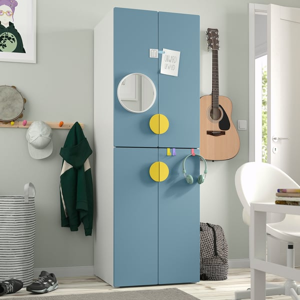 Blue SMÅSTAD wardrobe with yellow handles, mirror, & soft-close hinges next to door. Surrounded by a chair, guitar, & wall art. Compact & spacious.