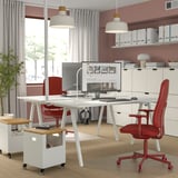 Modern home office with dual white desks, red chairs, hanging lamps, large monitor, and storage units. Chairs fit neatly under desks.