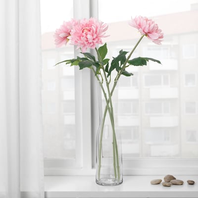 A clear glass vase holds two pink artificial flowers with green leaves, situated on a white window sill. The flowers appear realistic.