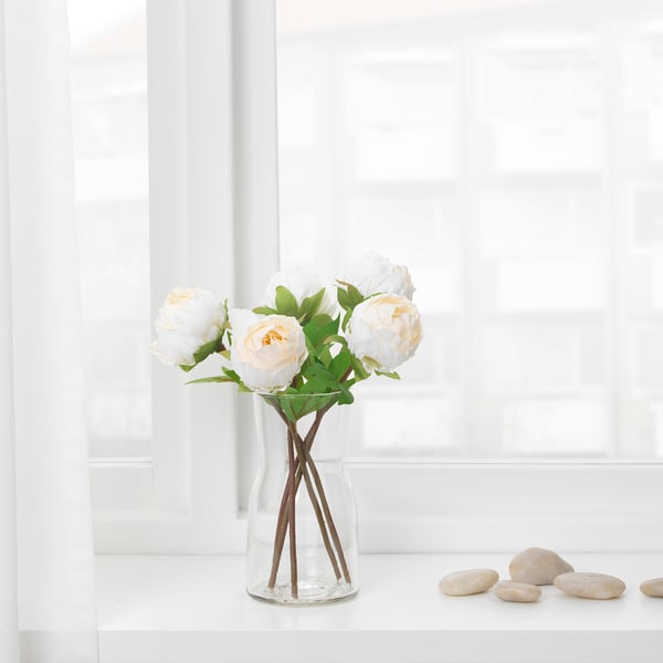 A clear glass vase with a stemmed cross holder displays white, peach flowers and green leaves. It sits on a white surface near a window.