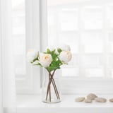 A clear glass vase with a stemmed cross holder displays white, peach flowers and green leaves. It sits on a white surface near a window.