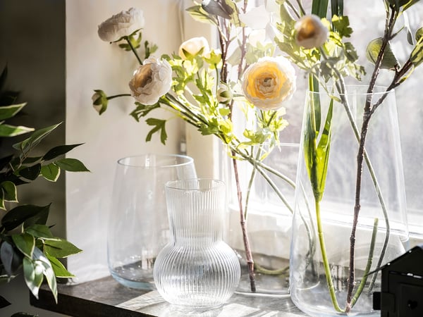 Three clear vases hold light-colored flowers and greenery, one vase with a ribbed texture, set on a windowsill.