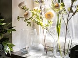 Three clear vases hold light-colored flowers and greenery, one vase with a ribbed texture, set on a windowsill.