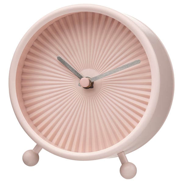 A pink, round SNOFSA clock with a sunburst design and silent quartz movement. It has a glass front, steel frame, and three spherical feet.