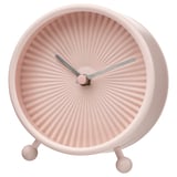 A pink, round SNOFSA clock with a sunburst design and silent quartz movement. It has a glass front, steel frame, and three spherical feet.
