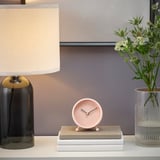 A modern clock sits on stacked books, beside a lamp and a vase with white flowers.