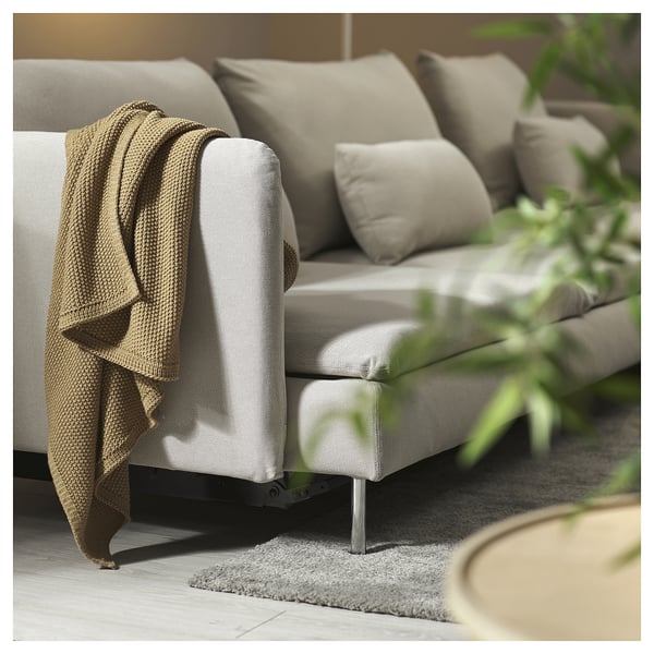 A neutral-toned, light grey SÖDERHAMN sofa bed with large cushions, a cosy blanket draped over one arm, and a small plant beside it.