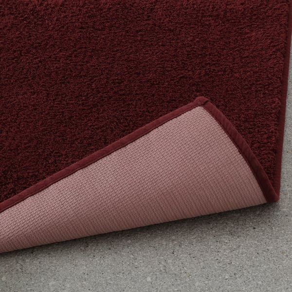 Burgundy bath mat with pivotable corner showing latex backing for secure grip on smooth surfaces.