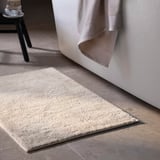 Beige SÖDERSJÖN bath mat on floor, soft cotton, rectangular with textured surface.