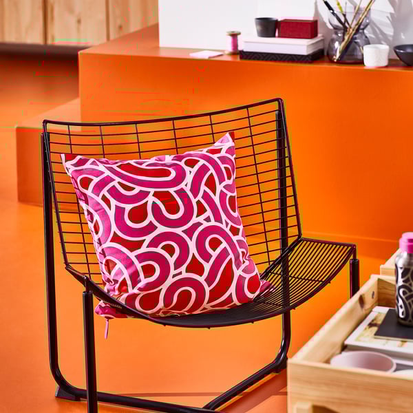 Black wire chair with bright pink cushion featuring abstract shapes. Sits against a matching orange wall.