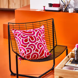 Black wire chair with bright pink cushion featuring abstract shapes. Sits against a matching orange wall.