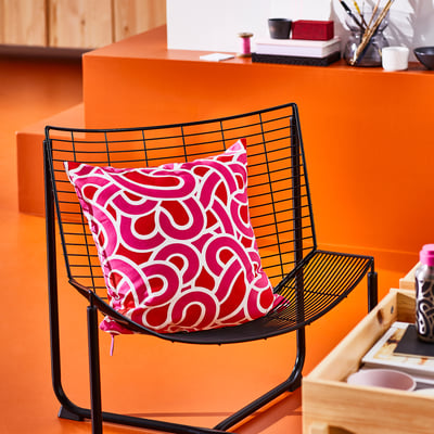 Black wire chair with bright pink cushion featuring abstract shapes. Sits against a matching orange wall.