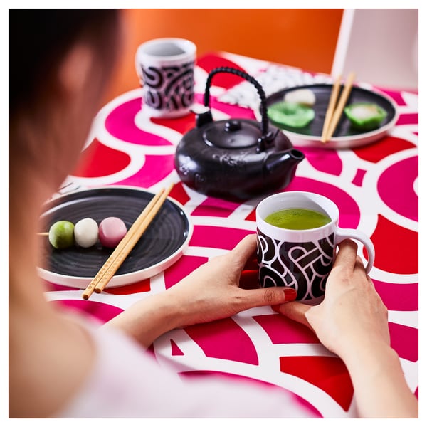 Person holding green tea mug on patterned table with sweets and teapot.