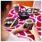 Person holding green tea mug on patterned table with sweets and teapot.